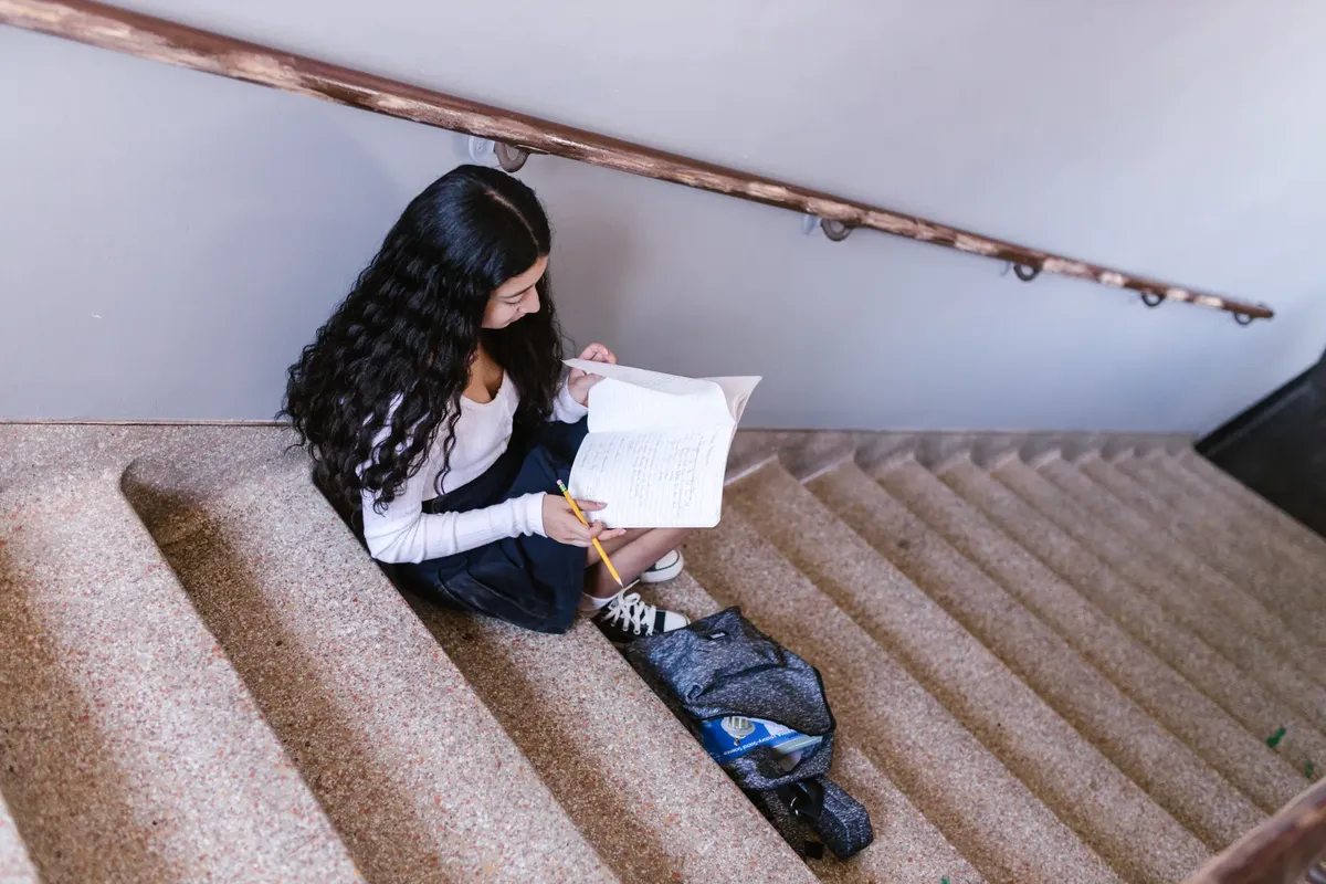 어린 여자는 학교 복도에서 구cret stairs에 앉아, 철봉과 노트북을 들고 공부하고 있다.