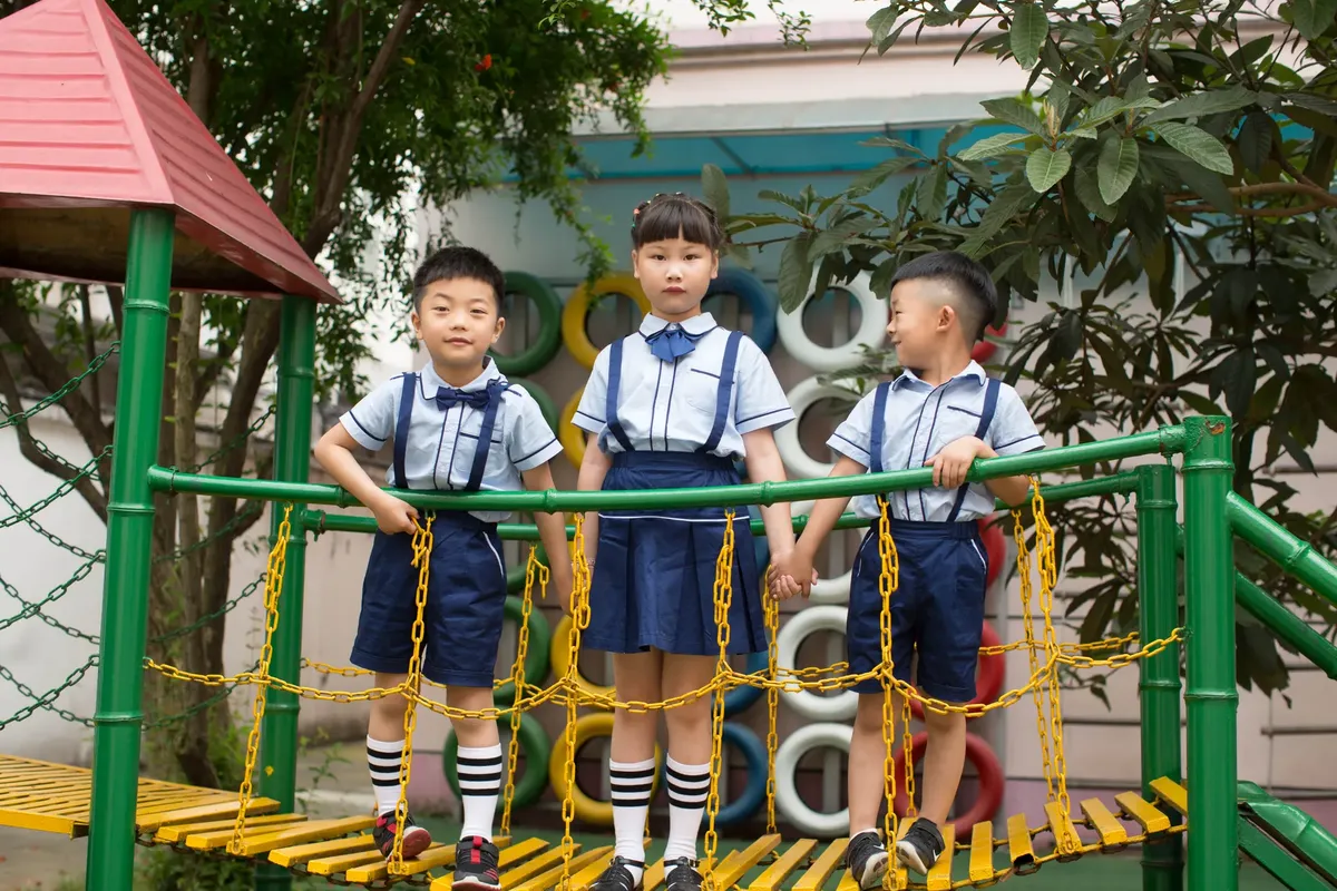 Three children in school uniforms standing on a playground bridge outdoors, holding hands.